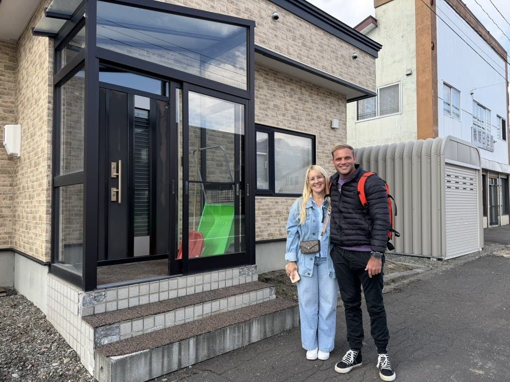 Michelle and Elliot standing arm in arm at the glass front porch of their house in Yubari, a snowboard rack visible inside the door.