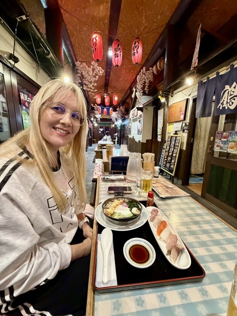 Michelle smiling at a table in a lantern-lit food hall, a tray of ramen and side dishes in front of her.