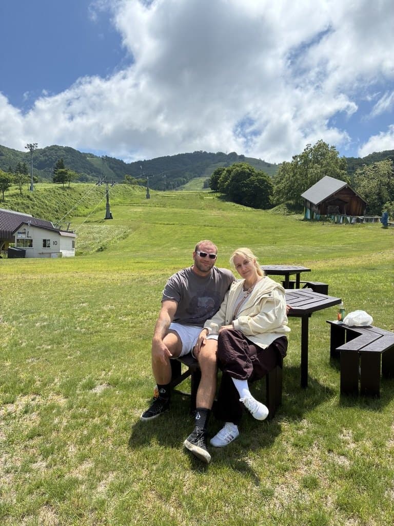 Elliot and Michelle sitting on a bench at the foot of a grassy ski slope in summer, chairlift towers running up the hill.