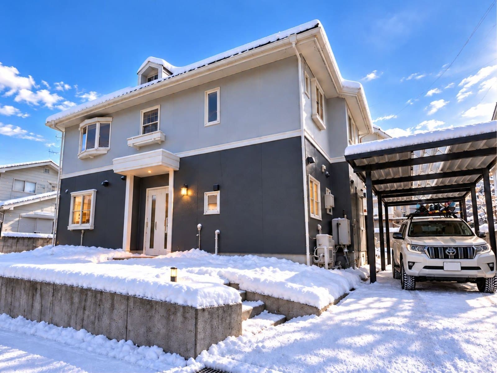 The Iizuna home in winter: a two-storey house in grey and white under a blue sky, snow on the garden and a car under the carport